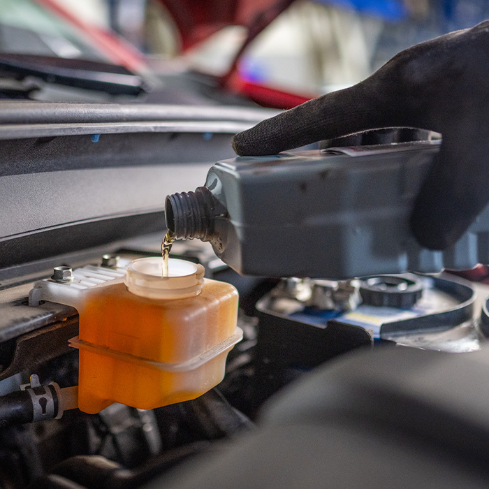 A mechanic is refilling brake fluid in a car engine bay, wearing protective gloves
