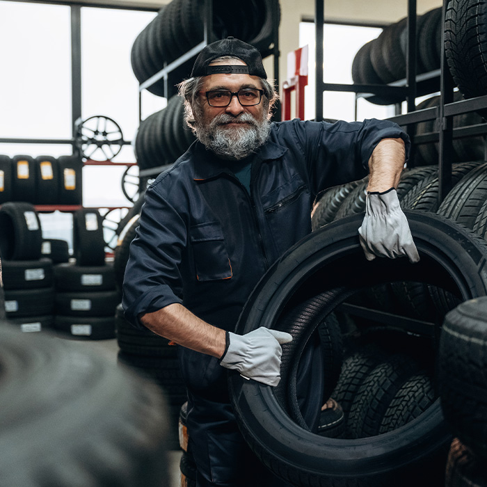 Senior male tire technician wearing blue coveralls holds a tire in a tire shop